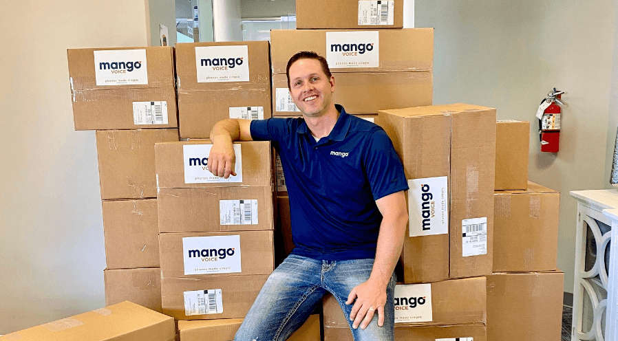 A Mango Voice team member smiling next to a large stack of Mango Voice shipping boxes in the office